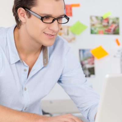 Creative worker. Cheerful young man in glasses working on computer and smiling while sitting at his working place